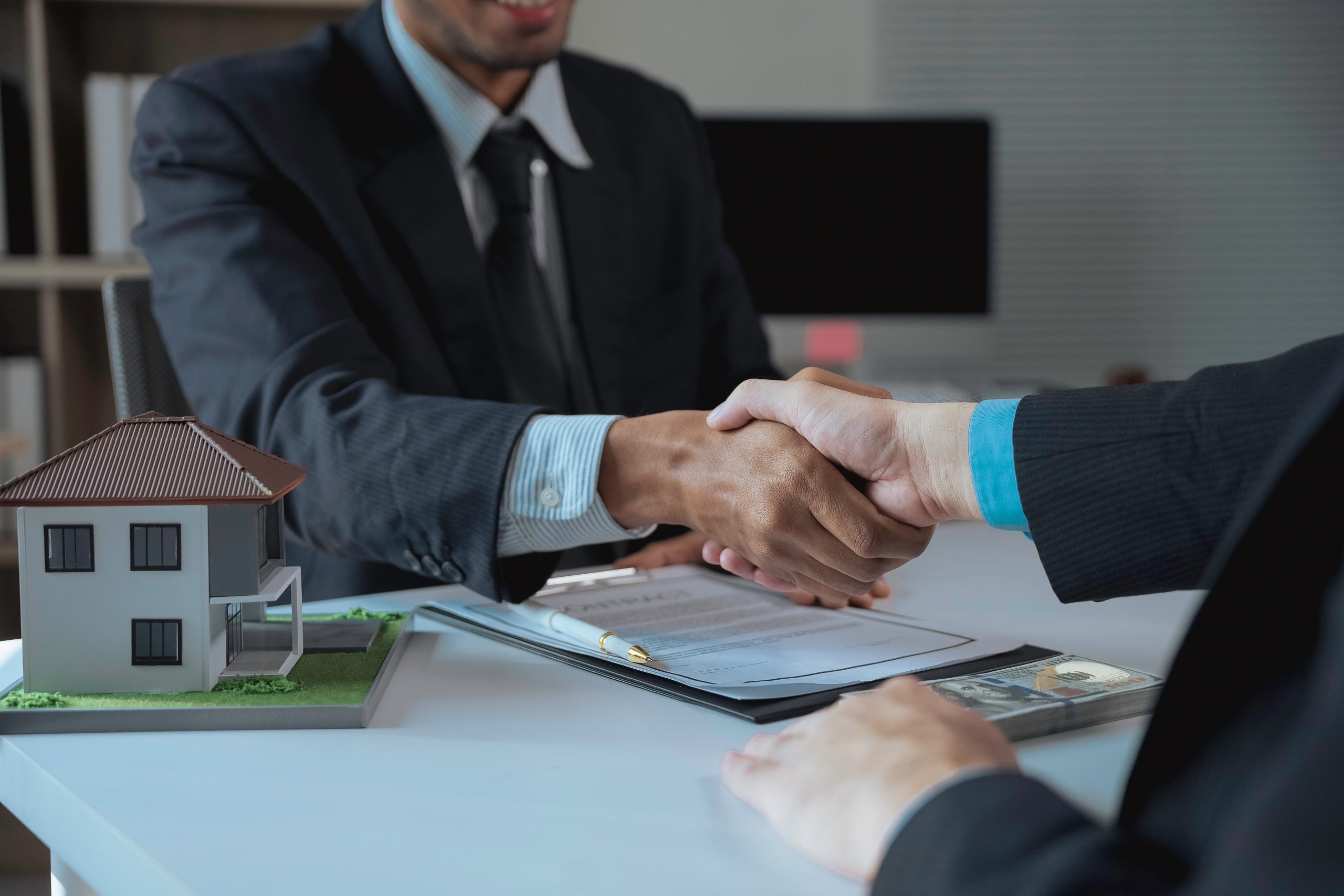 Real estate agent shaking hands with customer after successfully signing a contract for a new house and closing the deal in the office, miniature house model, contract and money on the table